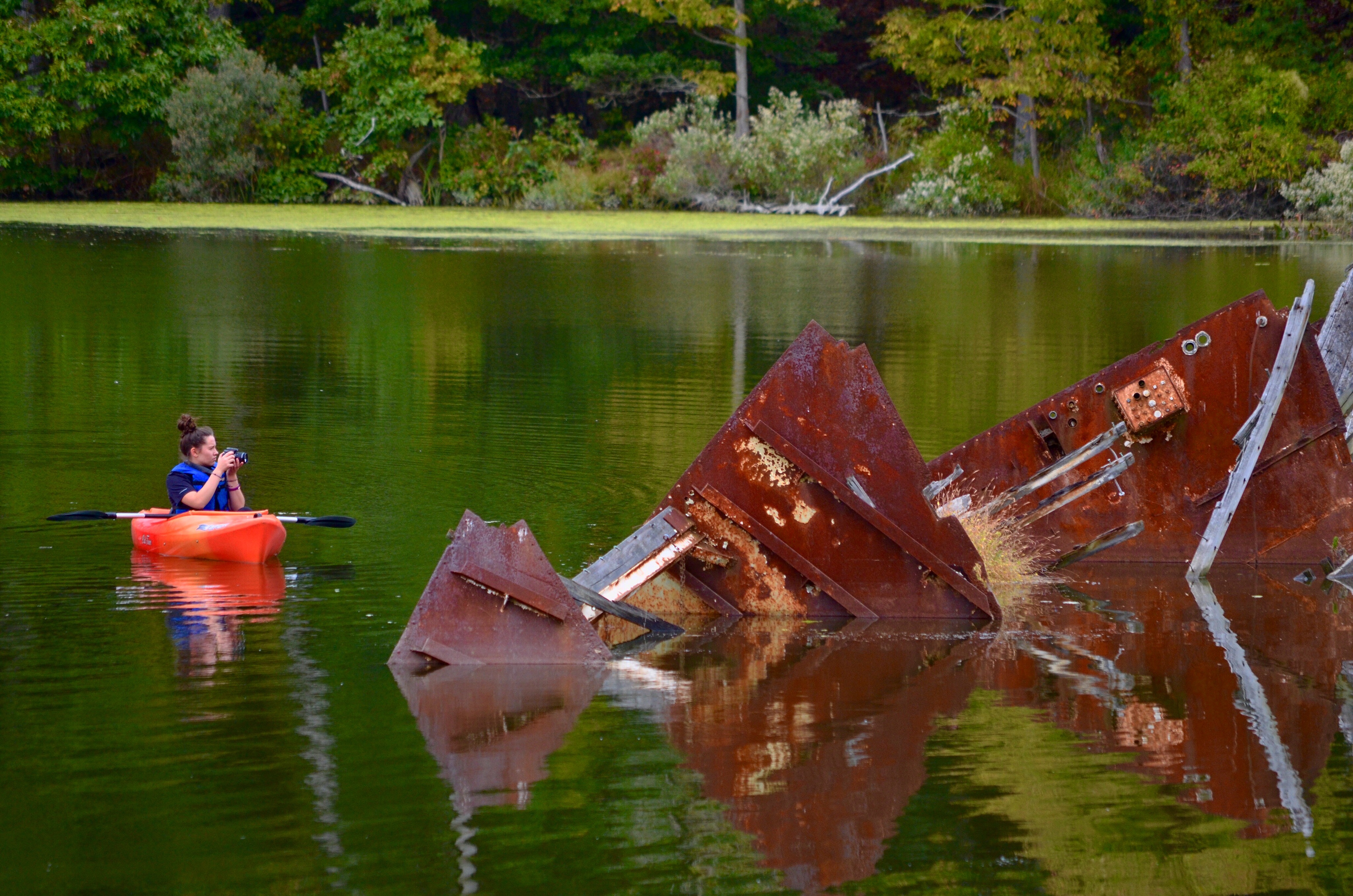 Lilly in a orange kayak capturing a rusty sunken barge with her camera.